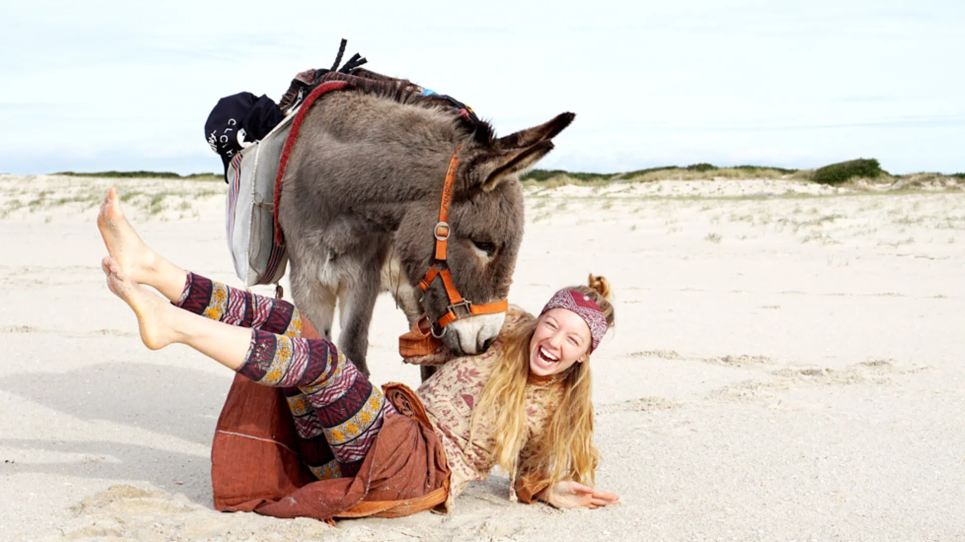 Eine Frau liegt lachend neben ihrem Esel im Sand.