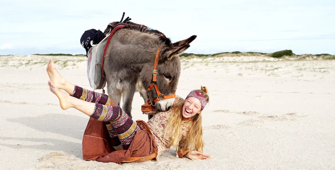 Eine Frau liegt lachend neben ihrem Esel im Sand.