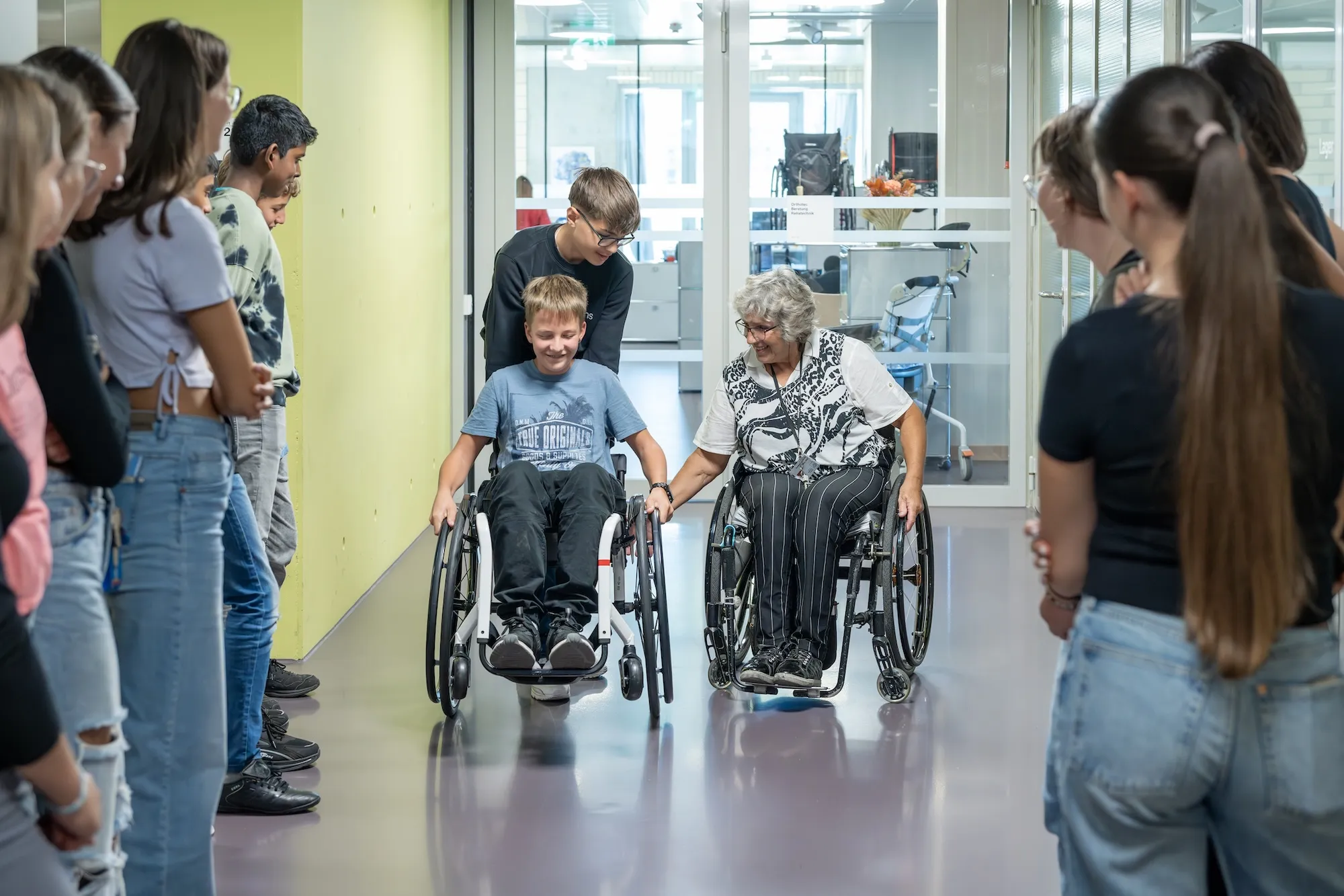 Rosa Zaugg zeigt als Gästebegleiterin für das Besuchszentrum der Schweizer Paraplegiker-Stiftung in einem Gang des SPZ einem Schüler, wie er sich im Rollstuhl fortbegwegen kann. Links und rechts säumen weitere Schüler der Klasse, die zu Besuch ist, den Gang und schauen zu.