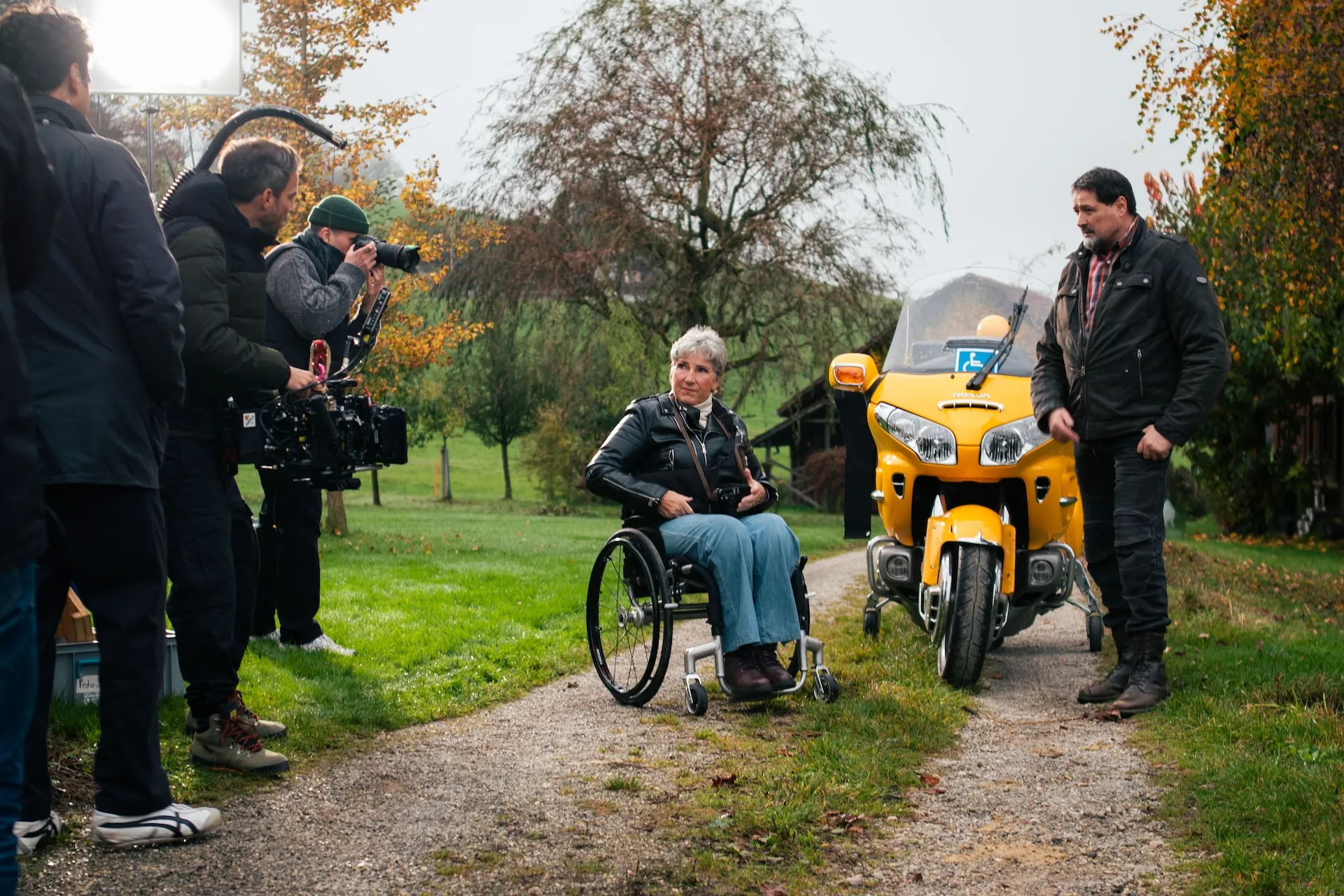 Rosa Zaugg im Rollstuhl und daneben ihr Partner Lukas auf einem Feldweg - im Hintergrund Lukas' Motorrad, eine gelbe Goldwing. Links von ihnen stehen Mitglieder der Filmcrew und machen erste Aufnahmen.