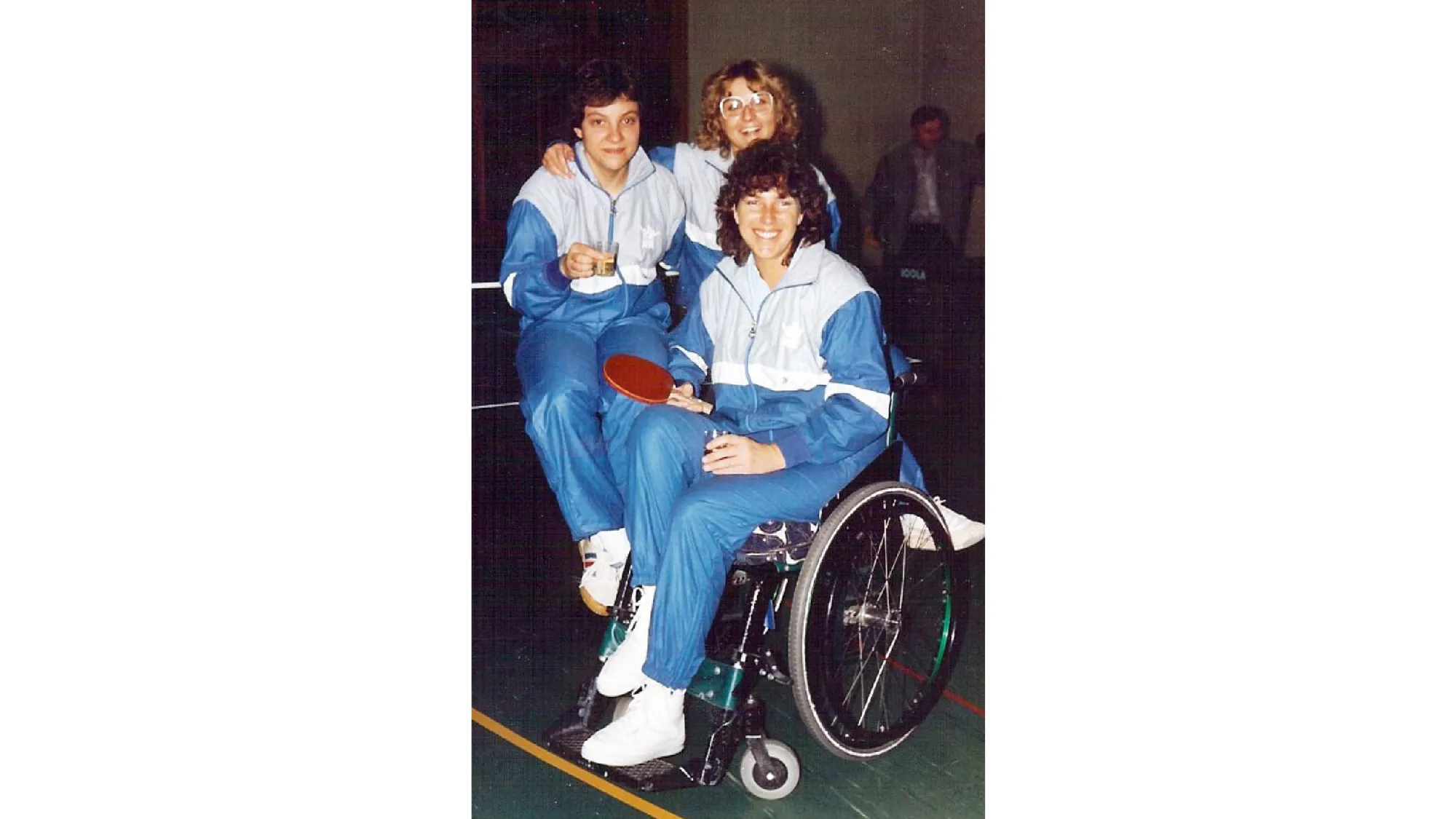 Rosa Zaugg im blau-weissen Trainings-Anzug im Rollstuhl. Sie hält einen Ping-Pong-Schläger in der Hand und lächelt in die Kamera. Hinter ihr stehen zwei weitere Frauen aus dem Tischtennisclub.