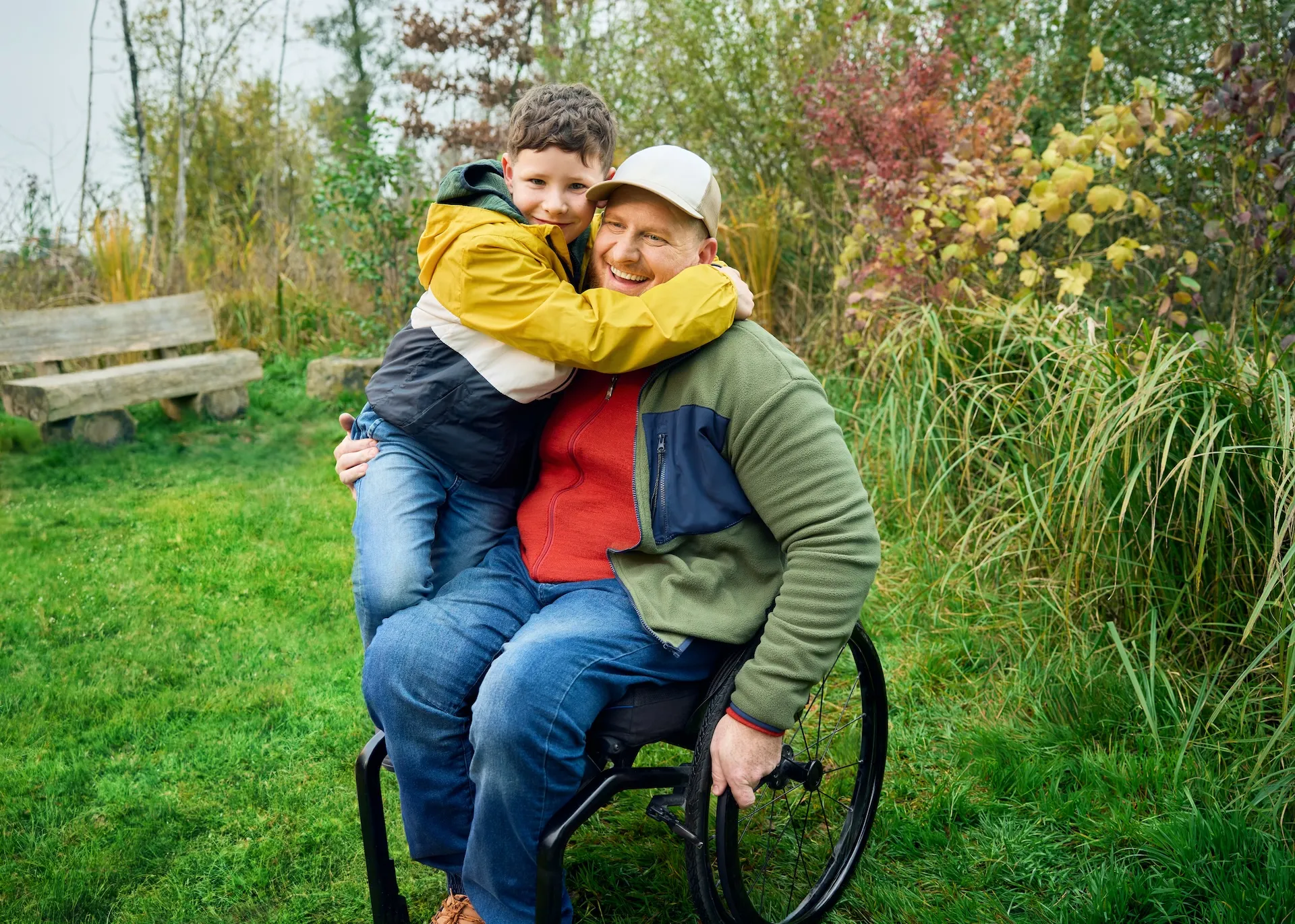 Heiri sitzt in seinem Rollstuhl auf einer Wiese und sein Sohn klettert ihm halbwegs auf den Schoss und umarmt seinen Vater ganz fest. Beide lächeln.