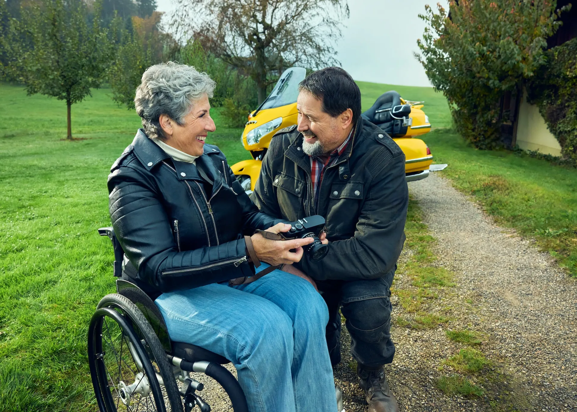 Rosa Zaugg sitzt in ihrem Rollstuhl auf einem Feldweg auf einem Bauernhof und blickt zu ihrem Partner Lukas hinüber, der sich neben ihr hingekniet hat. Sie zeigt ihm eine Kamera. Im Hintergrund ist seine gelbe Goldwing zu sehen.