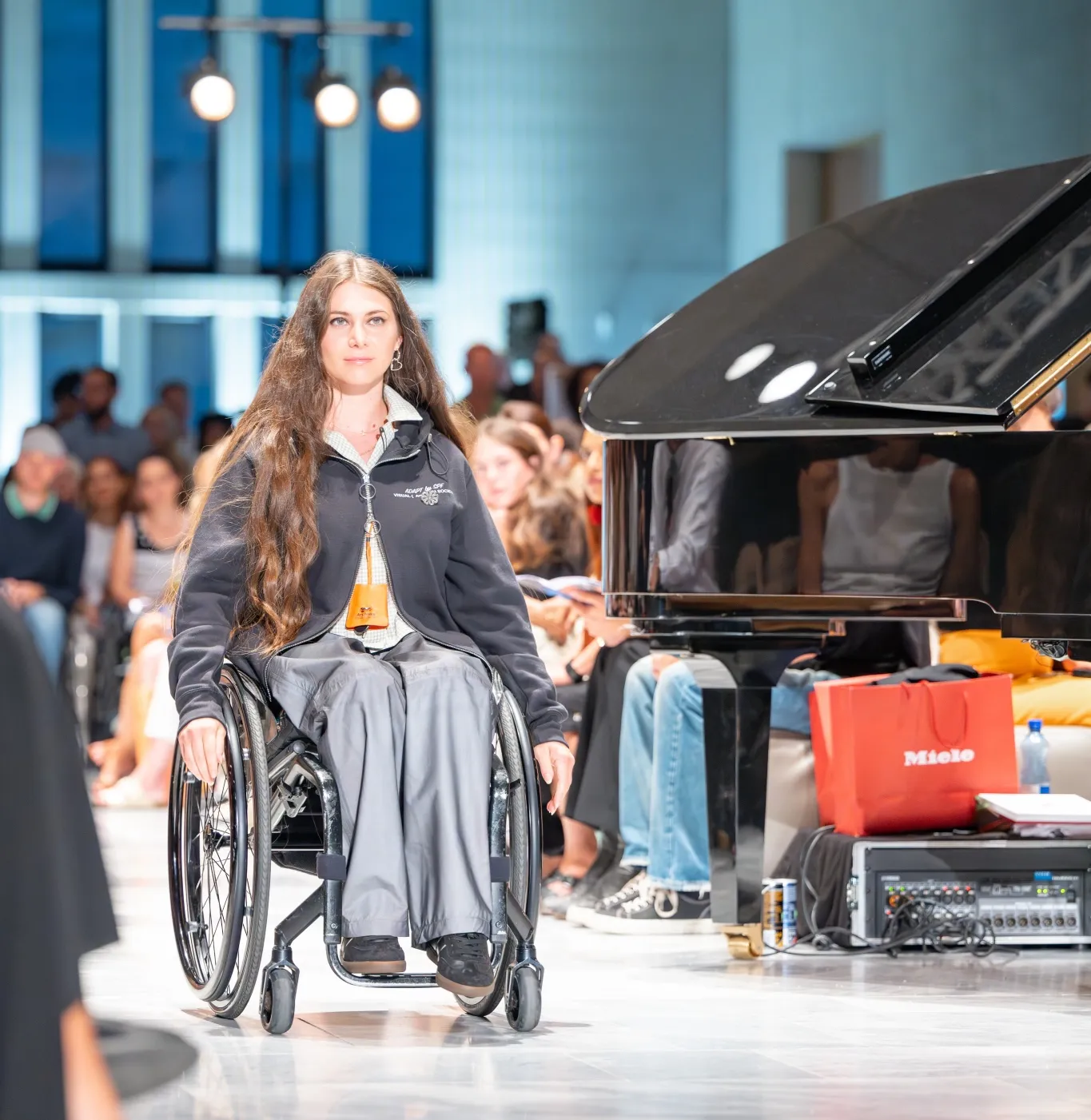 Chiara Schlatter rollt auf dem Catwalk in ihrem inklusiven Outfit an den Zuschauern vorbei. Sie trägt eine schwarze Jacke und eine silbrige, lockere Hose. Neben dem Catwalk ist ein Klavier-Flügel.