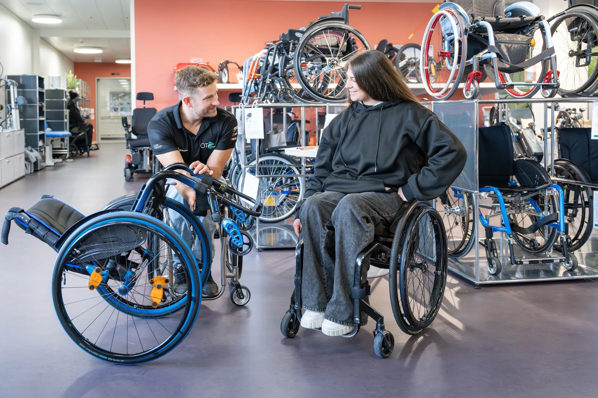 Chiara sitzt vor einem Regal voller Rollstühle in ihrem Rollstuhl, neben ihr kniet ein Mitarbeiter der Orthotec am Boden und hält einen Rollstuhl gekippt, um Chiara daran etwas zu zeigen.