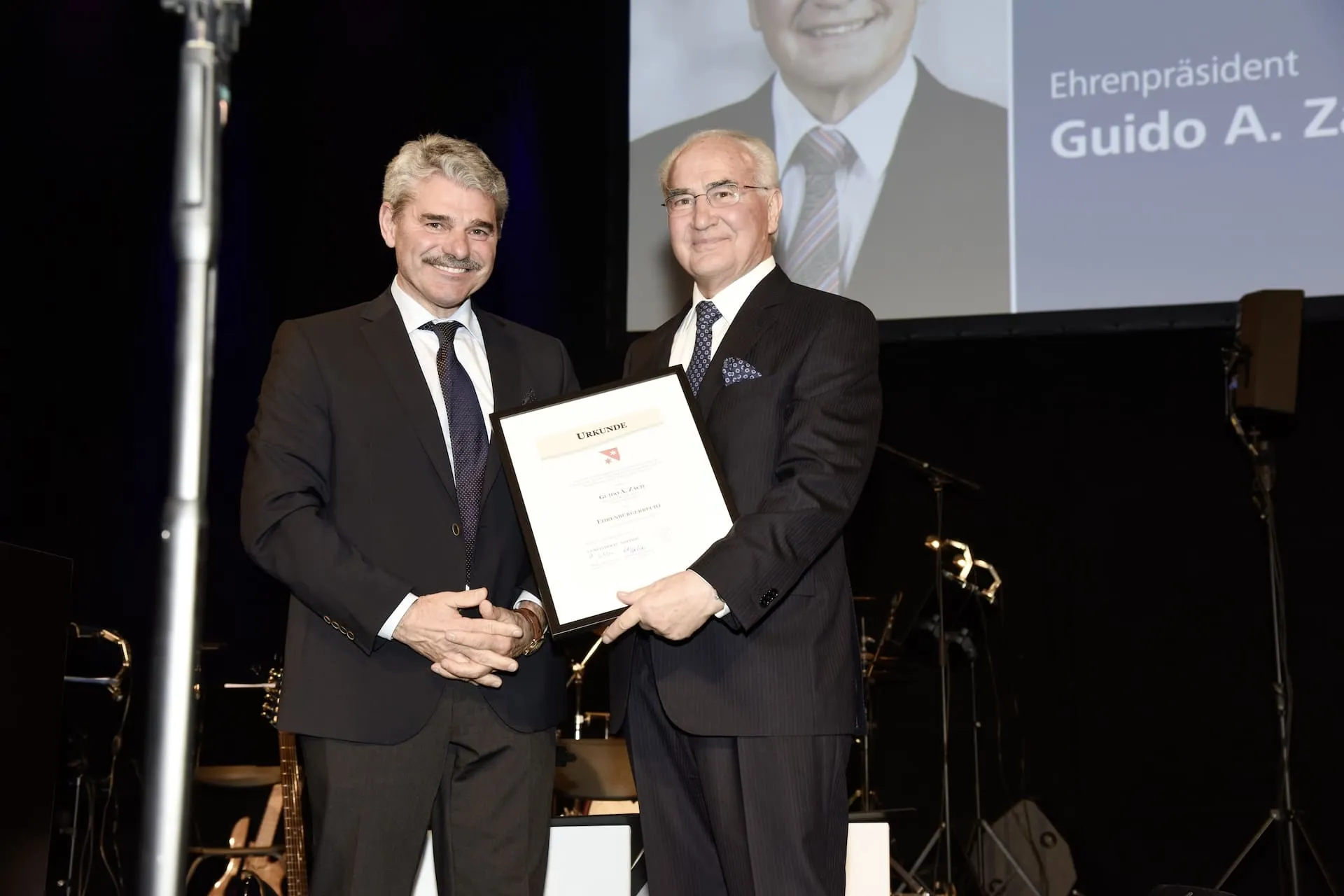 Guido A. Zäch steht neben Nottwiler Gemeinderatspräsident Walter Steffen auf einer Bühne vor schwarzem Hintergrund und einer Projektsionsfläche mit Foto von Guido A. Zäch. Er hält ein Dokument mit der Ehrenbürgerschaft in der Hand. Beide lächeln in die Kamera.