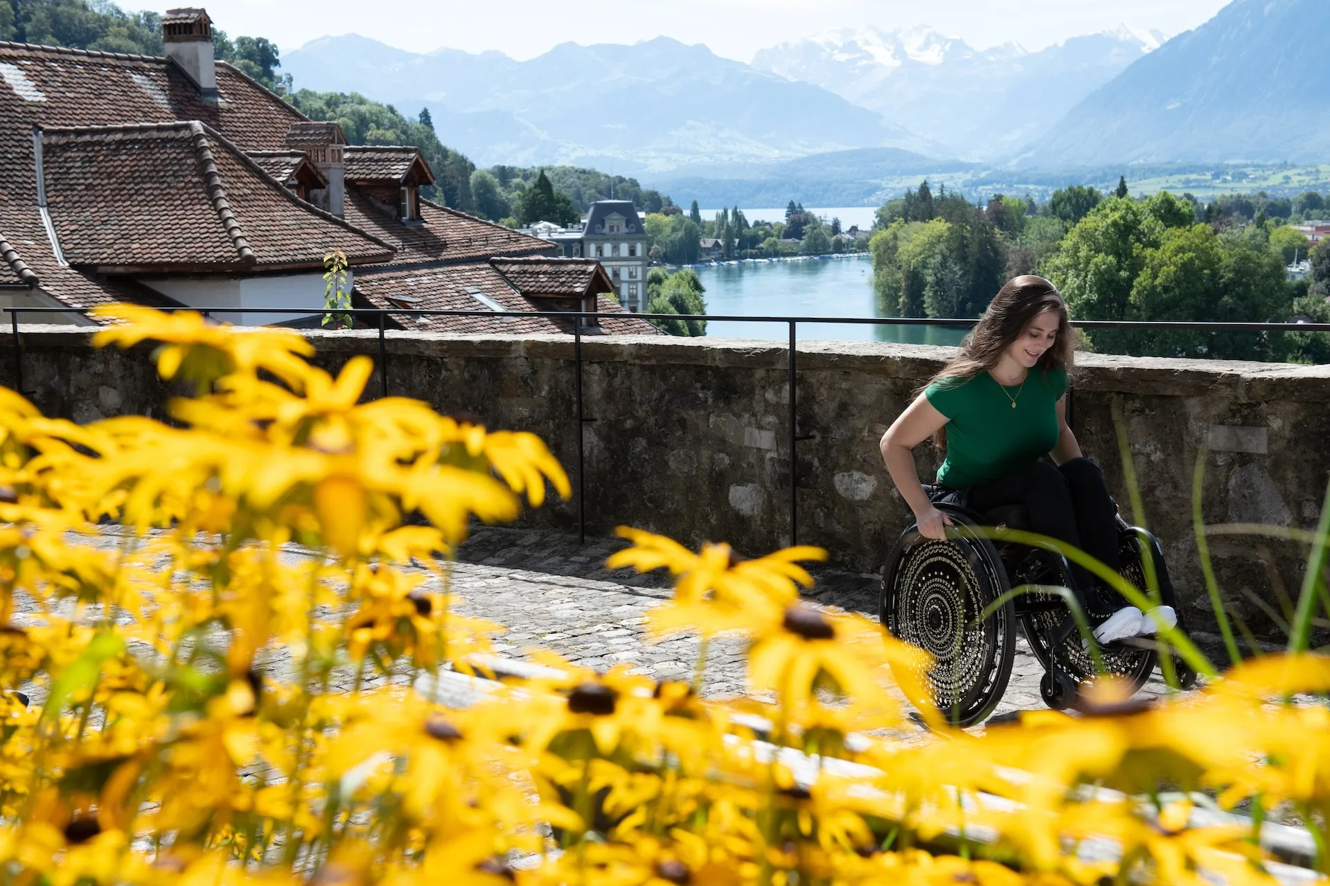 Chiara rollt über eine Brücke, im Vordergrund sind gelbe Blume zu sehen und im Hintergrund ein Fluss und Hausdächer sowie ein Bergpanorama.