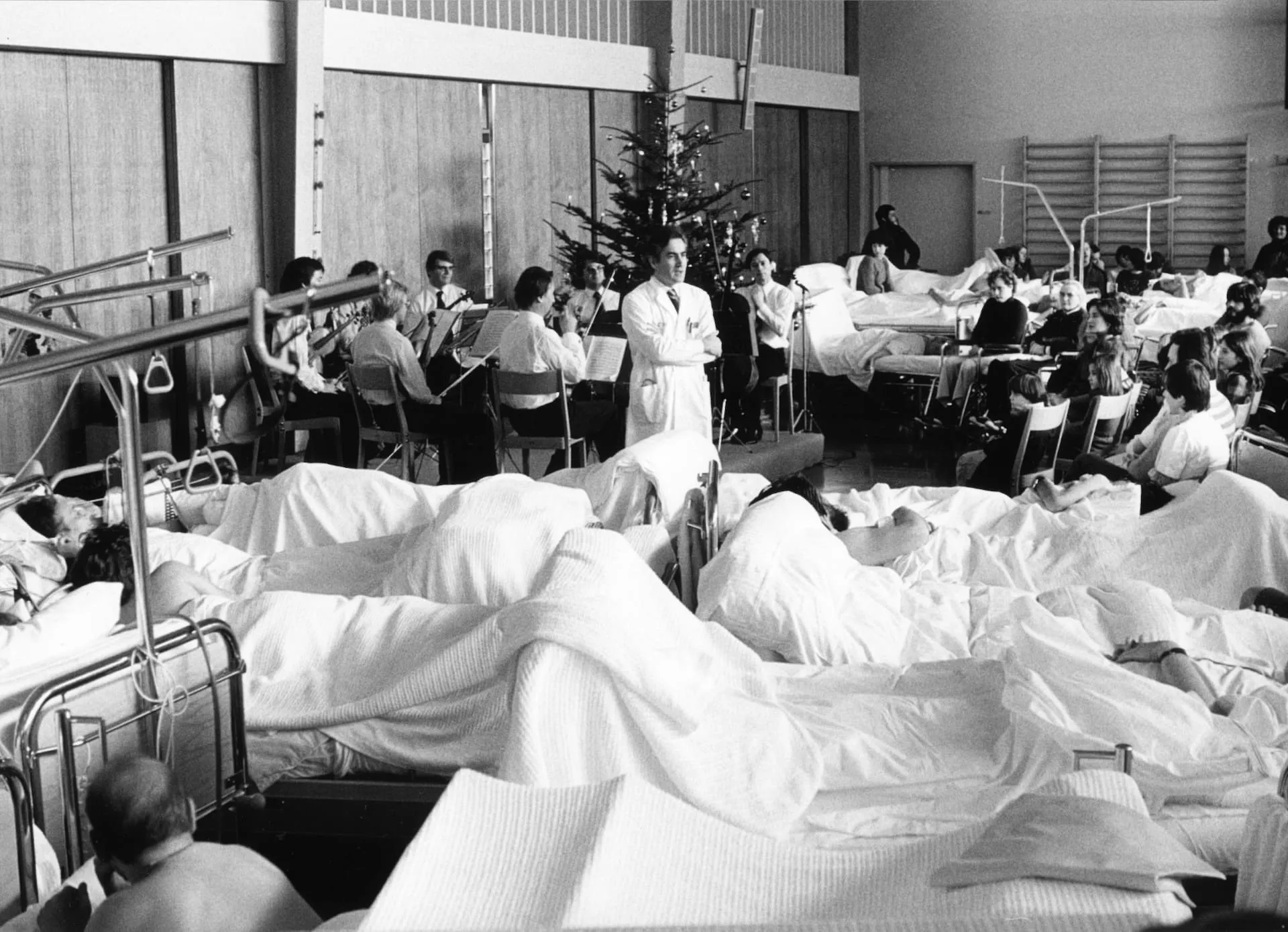 Schwarz-Weiss-Foto: Im Vordergrund sind viele Betten mit Patienten zu sehen, im Hintergrund ein Weihnachtsbaum und ein Orchester, das ein Konzert gibt. Guido A. Zäch steht in der Mitte des Fotos.