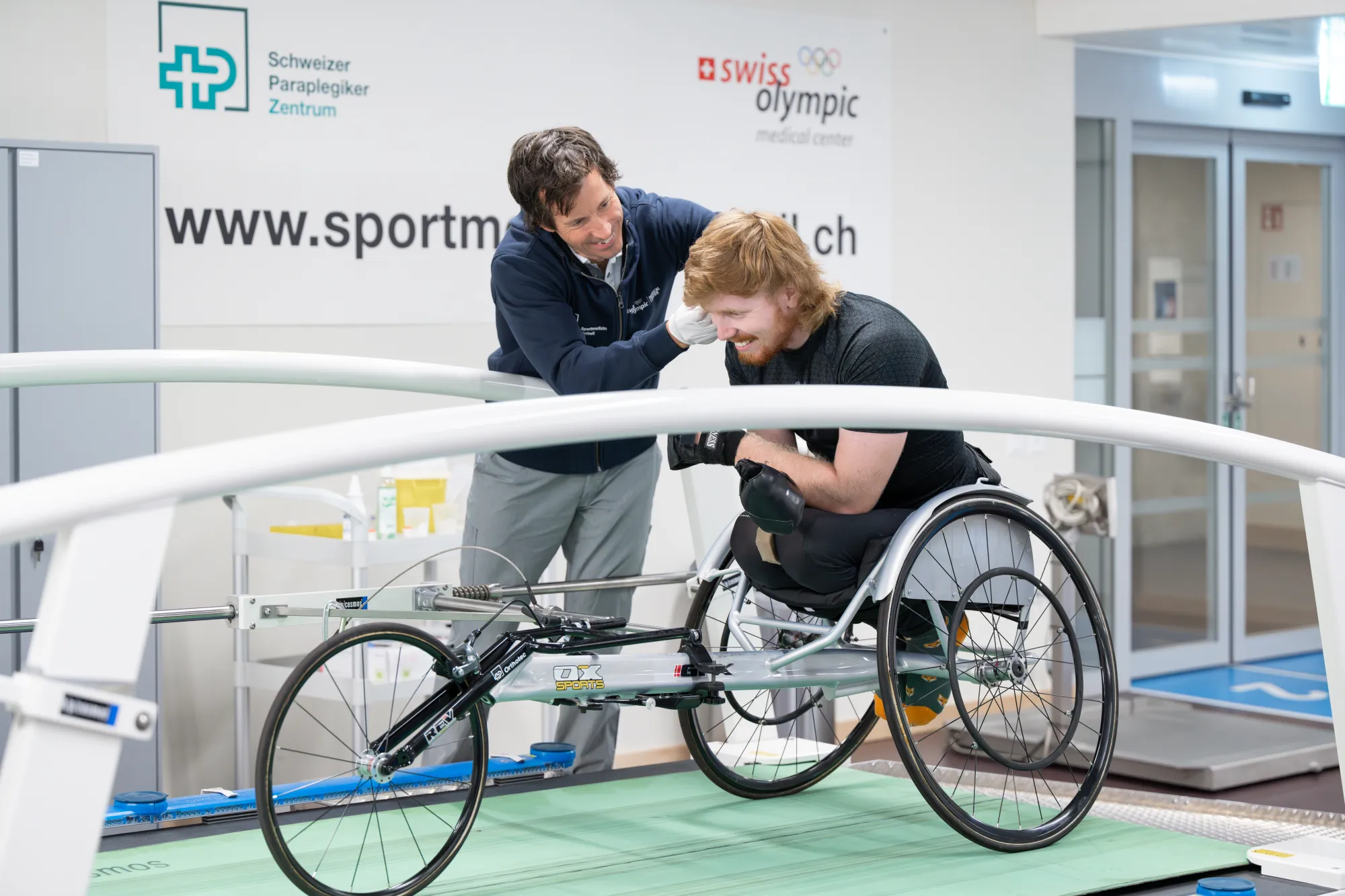 Tim Harder auf einem Rennrollstuhl auf einem grünen Laufband. Daneben steht ein Mann, er ihn an der Schulter berührt und anschaut. Die beiden sind im Sportmedizinzentrum des Schweizer Paraplegiker-Zentrums.