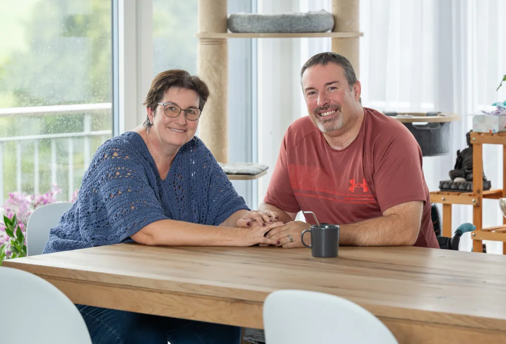 Pierre-Alain Tercier und Mireille Schafer sitzen am Tisch, halten sich an den Händen und lächeln in die Kamera.